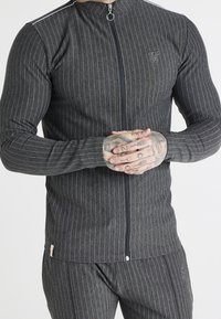 Grårandig zip-up jacka i texturerat tyg, med hög krage, ribbade muddar och broderad logotyp på bröstet.