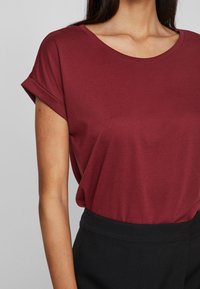 Μπλούζα σε μπορντό απόχρωση με στρογγυλή λαιμόκοψη και κοντά μανίκια που έχουν τυλιχθεί, από μαλακό ύφασμα. Συνδυασμένη με μαύρο παντελόνι ψηλής μέσης.