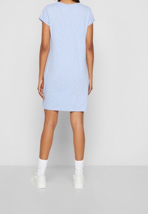 Woman standing, wearing a light blue short-sleeve dress, white socks, and white sneakers, facing away against a plain gray background.