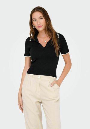 Polo nero a maniche corte con coste e scollatura a V, con strisce bianche di accentuazione sul colletto e sulle maniche. Abbinato a pantaloni beige chiaro.
