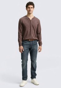 Herren-Henley-Shirt in Weinrot mit langem Arm und leichter Textur, kombiniert mit blauen Jeans und weißen Turnschuhen, vor hellem Hintergrund stehend.