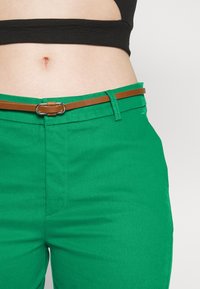 Nagie brzuch w czarnym topie crop, wysokiej talii zielone spodnie z pętlami na pasek oraz cienkim brązowym paskiem ze srebrną klamrą.