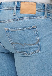 Svetlo modré džínsy s zadnou vreckom, ktoré má žlté šitie. Na opasku je hnedá kožená nášivka s nápisom "JACK & JONES."
