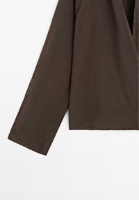 Bruin kledingstuk met lange mouwen, een losse pasvorm en zichtbare naaddetails op de mouw en zoom, afgebeeld tegen een witte achtergrond.