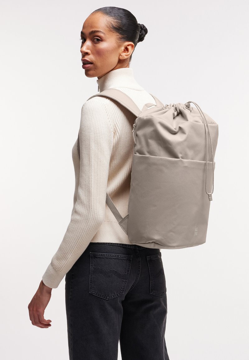 Kvinde iført beige rullekravetrøje og sorte jeans bærer stor beige rygsæk med snøre over den ene skulder mod hvid baggrund.