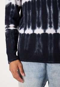 Sweatshirt tie-dye bleu marine et blanc avec une finition texturée, à manches longues et col rond, associé à un denim bleu clair.