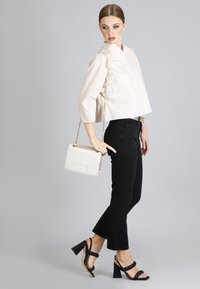 Camicia beige chiaro tagliata con dettagli in bottoni, abbinata a jeans neri a zampa d'elefante e sandali neri con tacco intrecciato. Borsa bianca con tracolla in catena dorata.