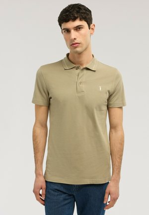 Junger Mann mit dunklem lockigem Haar, der ein beiges kurzärmeliges Poloshirt und blaue Jeans trägt und vor einem schlichten hellen Hintergrund steht.