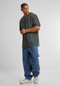 Dark green cotton t-shirt with a crew neck and short sleeves, featuring black printed text. Paired with light blue baggy denim jeans and beige sneakers.