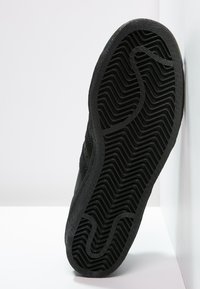 Mustan urheilukengän pohja, jossa on sahalaitakuviot, tekstuuroitu kumimateriaali ja hieman kohotettu reuna. Näkyvissä on kulumisen merkkejä ja pitoon liittyvää muotoilua.