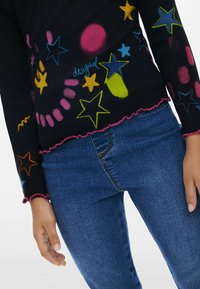 Schwarzes geripptes Langarmoberteil mit bunten Stern- und Wirbelmustern, einem ausgefransten rosa Saum und Bündchen, kombiniert mit blauen Jeans.