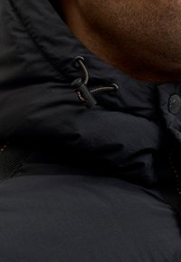 Chaqueta acolchada negra con capucha que presenta un tejido texturizado, cordones ajustables y un detalle de costura en naranja contrastante en la costura.