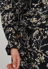 Musta kukkakuosiset blouse, jossa on valkoisia ja kultaisia kasvipainatuksia, kimaroidut hihat ja nyörityksellä varustetut yksityiskohdat. Teksturoitu kangas näkyy selvästi.