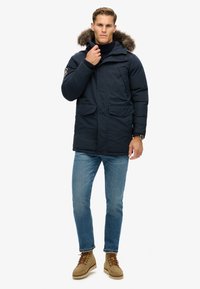 Marineblaue Winterjacke mit einem fellgefütterten Kapuzen, Reißverschluss vorne, zwei Fronttaschen, getragen über einem marineblauen Pullover und kombiniert mit hellblauen Jeans und beigen Stiefeln.