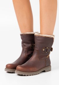 Bottines en cuir marron avec doublure en fausse fourrure, détail de boucle sur le côté, et semelles en caoutchouc texturé avec un motif de crampons.