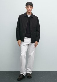 Young man wearing a black jacket, black sweater, white pants, and dark brown shoes standing against a plain light gray background.