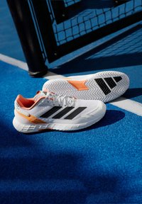 Λευκά αθλητικά παπούτσια με μαύρες ρίγες και πορτοκαλί λεπτομέρειες, με πλέγμα υφάσματος και καουτσούκ στο κάτω μέρος, και έντονο σχέδιο ζιγκ-ζαγκ.