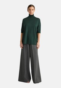 Vrouw in een donkergroene coltrui met ellebooglange mouwen en wijde grijze broek, staande tegen een effen achtergrond.