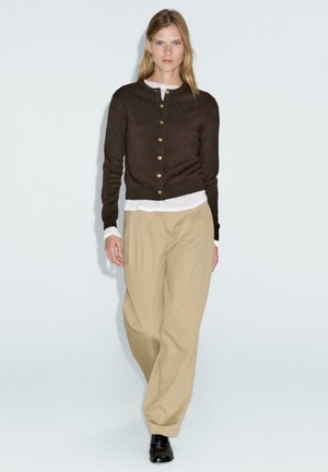 Brauner Cardigan mit goldenen Knöpfen, getragen über einem weißen Oberteil, kombiniert mit beigen weiten Hosen. Der Stoff scheint weich und hat eine lässige Passform.