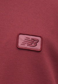 Burgundia tekstiil, millel on tekstureeritud pind, ja mis sisaldab roosa nelinurkse plaastri, millel on New Balance'i logo.