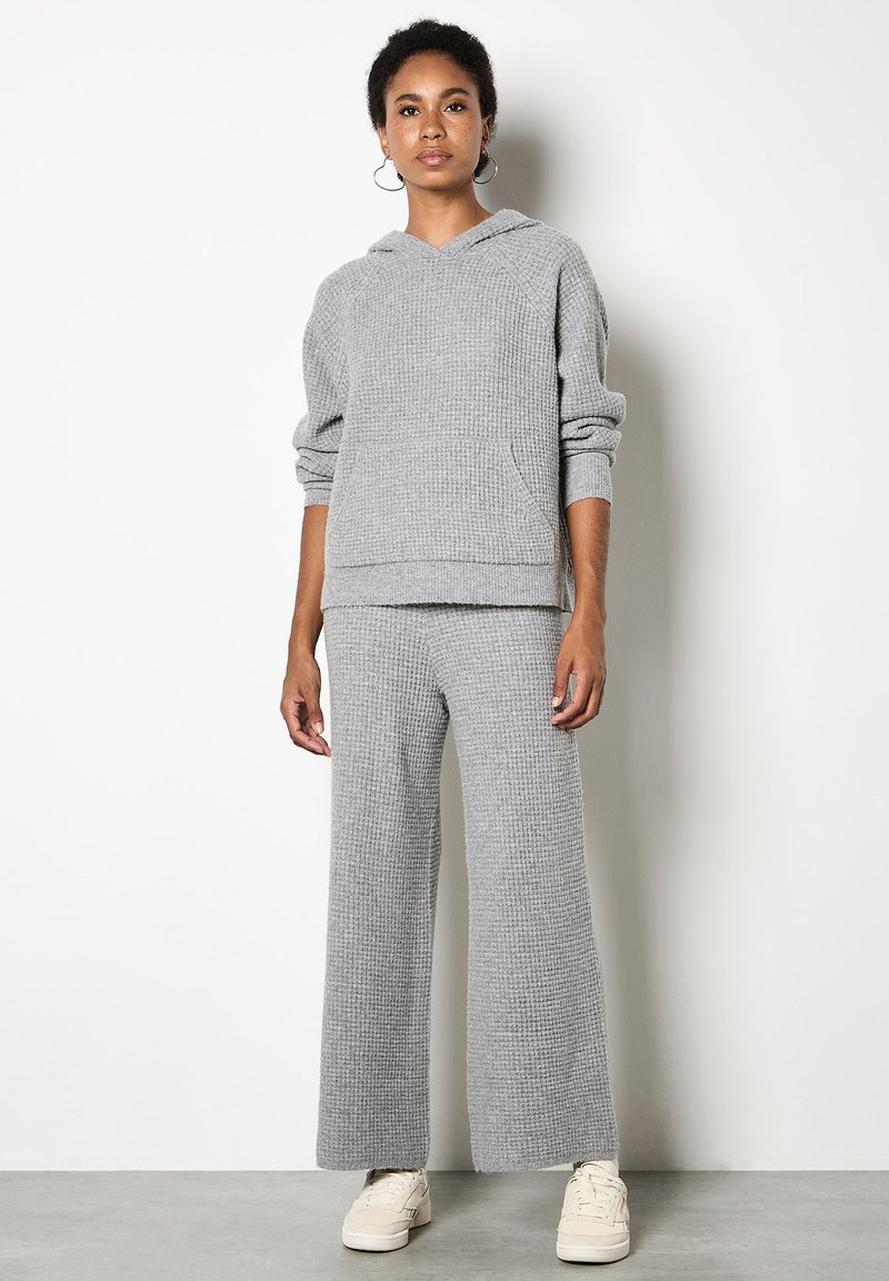 Szara dzianinowa bluza z kapturem z kieszenią oraz szerokimi spodniami w komplecie. Miękka tekstura, luźny krój i swobodny design. Białe sneakersy dopełniają stylizację.