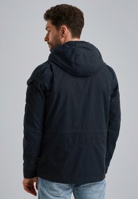 Giacca blu navy con cappuccio, caratterizzata da un tessuto liscio e testurizzato, tasche laterali e spalle strutturate. La vista posteriore mostra una vestibilità rilassata.