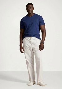 Άνδρας φορώντας ένα ναυτικό μπλε κοντομάνικο μπλουζάκι με V λαιμόκοψη, ανοιχτό μπεζ παντελόνι, λευκά αθλητικά παπούτσια και ρολόι καρπού, στέκεται μπροστά από ένα απλό φόντο.