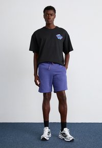 T-shirt noir oversized avec des détails graphiques, associé à un short violet et des baskets blanches. Fond uni, le modèle se tient debout.