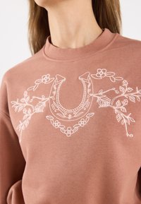 Rosa genser med hvit broderte blomster og hestesko-design på forsiden, med høy hals og myk tekstur.