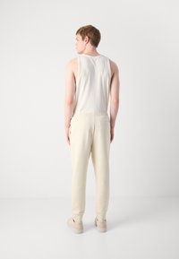 Jonge man die met zijn rug naar de camera staat, gekleed in een geribbelde witte mouwloze top, crèmekleurige joggers en beige sneakers, tegen een effen achtergrond.
