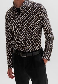 Zwart patroonhemd met een geometrisch ontwerp in wit en bruin, voorzien van een kraag en een knoopsluiting aan de voorkant, gecombineerd met zwarte broek en een riem.