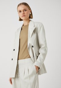 Světle šedý oblekový blazer s jemným proužkem, se dvěma předními kapsami a černými knoflíky, spárovaný s krémovým upnutým topem a odpovídajícími upravenými kalhotami.