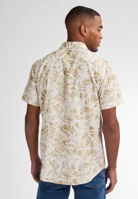 Camisa de botones de manga corta en tejido beige con un patrón de hojas tropicales doradas, cuello estándar y dobladillo curvado.