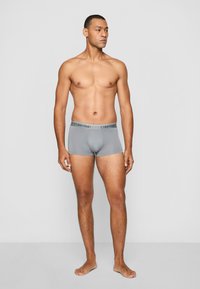 Graue Boxershorts mit passgenauer Schnittform, Stoffstruktur und elastischem Taillenbund mit dem Calvin Klein-Logo. Das Model steht barfuß.