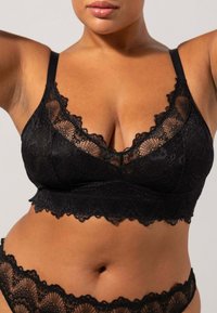 Mustavillainen pitsinen bralette, jossa on aaltoilevat reunat, säädettävät olkaimet ja V-mallinen kaula-aukko. Pehmeä pinta, jossa on monimutkaisia kukkakuoseja.
