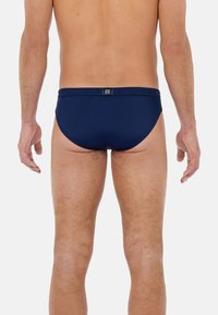 Námořnické pánské plavky s hladkým povrchem, nízkým střihem a elastickým pasem; na zadní straně má malou značkovou nálepku.