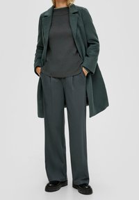 Manteau en laine vert foncé sur une chemise à manches longues gris foncé, associé à un pantalon large gris et des chaussures à plateforme noires chunky.