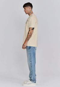 Beige Baumwoll-T-Shirt mit kurzen Ärmeln, kombiniert mit hellblauen Jeans. Das Model hat Tattoos an den Armen und trägt weiße Sneaker.