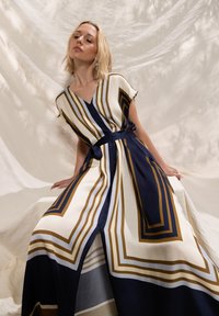 Abito fluente in beige, blu navy e oro con strisce geometriche. Presenta una scollatura a V e maniche corte, chiuso con una cintura abbinata. Tessuto dalla texture liscia.