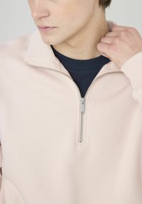 Sweatshirt rose clair avec un col à demi-fermeture éclair, présentant une couche intérieure bleu marine. Texture lisse, design minimaliste et détail de fermeture éclair en argent.