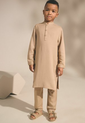 Junge steht, trägt eine beige traditionelle lange Tunika, passende Hose und Sandalen vor einem einfarbigen hellen Hintergrund mit Blockformen in der Nähe.