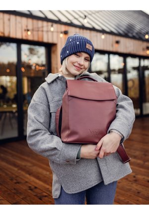 Maroon leather backpack with a flap, front logo, and red strap, held by a person wearing a grey coat and navy beanie in a wooden setting.