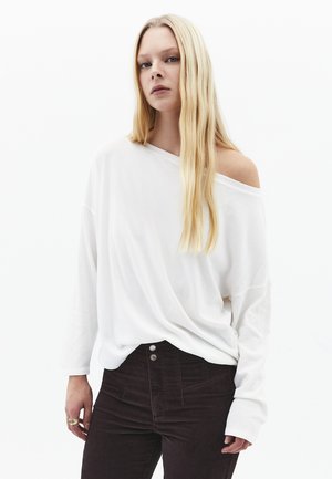 Mujer rubia con cabello largo, vistiendo un top blanco suelto que deja los hombros al descubierto y pantalones negros, posando frente a un fondo blanco.