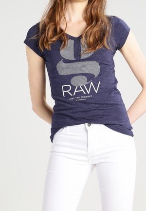 Femme portant un t-shirt graphique bleu marine avec le texte "RAW" et un pantalon blanc, debout les mains derrière le dos sur un fond clair uni.