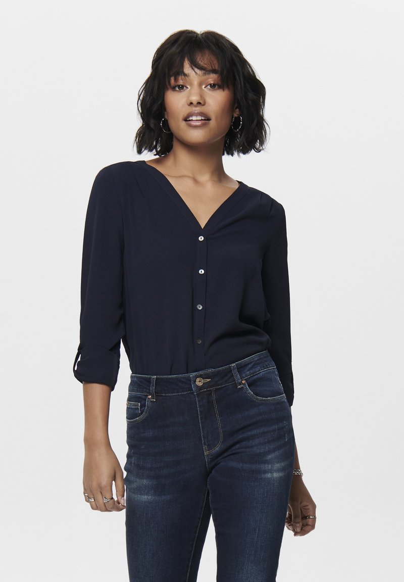 Blusa navy a maniche lunghe con scollo a V e chiusura a bottone. Tessuto morbido con una trama delicata. Abbinata a jeans scuri attillati, dotati di tasche classiche.