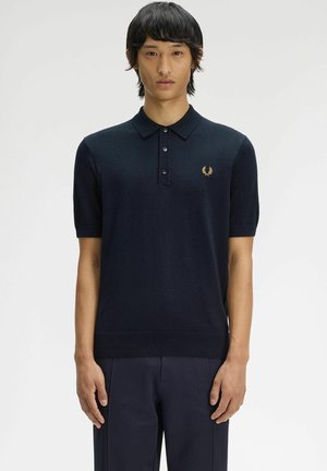 Uomo che indossa una polo blu navy a maniche corte con un piccolo logo di alloro dorato sul petto, in piedi davanti a uno sfondo bianco semplice.