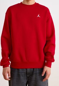 Person wearing a red crewneck sweatshirt with a small white Jumpman logo on the left chest and dark gray jeans.