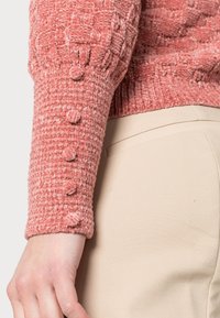 Pull rose tricoté avec un motif texturé, doté d'un poignet côtelé orné de quatre boutons recouverts de tissu. Associé à un pantalon beige.