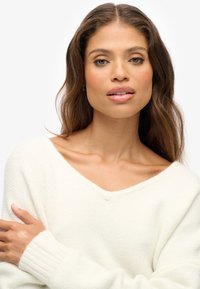 Crèmekleurige gebreide trui met een brede V-hals, geribbelde manchetten en een losse pasvorm, voorzien van een zachte textuur en subtiele stikdetails.