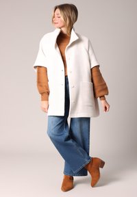 Weißer strukturierter Cardigan mit kurzen Ärmeln, mit Stehkragen und vorderen Knöpfen, getragen über einem braunen Pullover und blauen weit geschnittenen Jeans mit braunen Stiefeln.